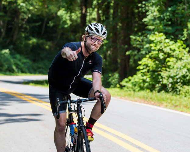 Asheville resident on bike gives a thumbs up while biking uphill on sunlit paved road